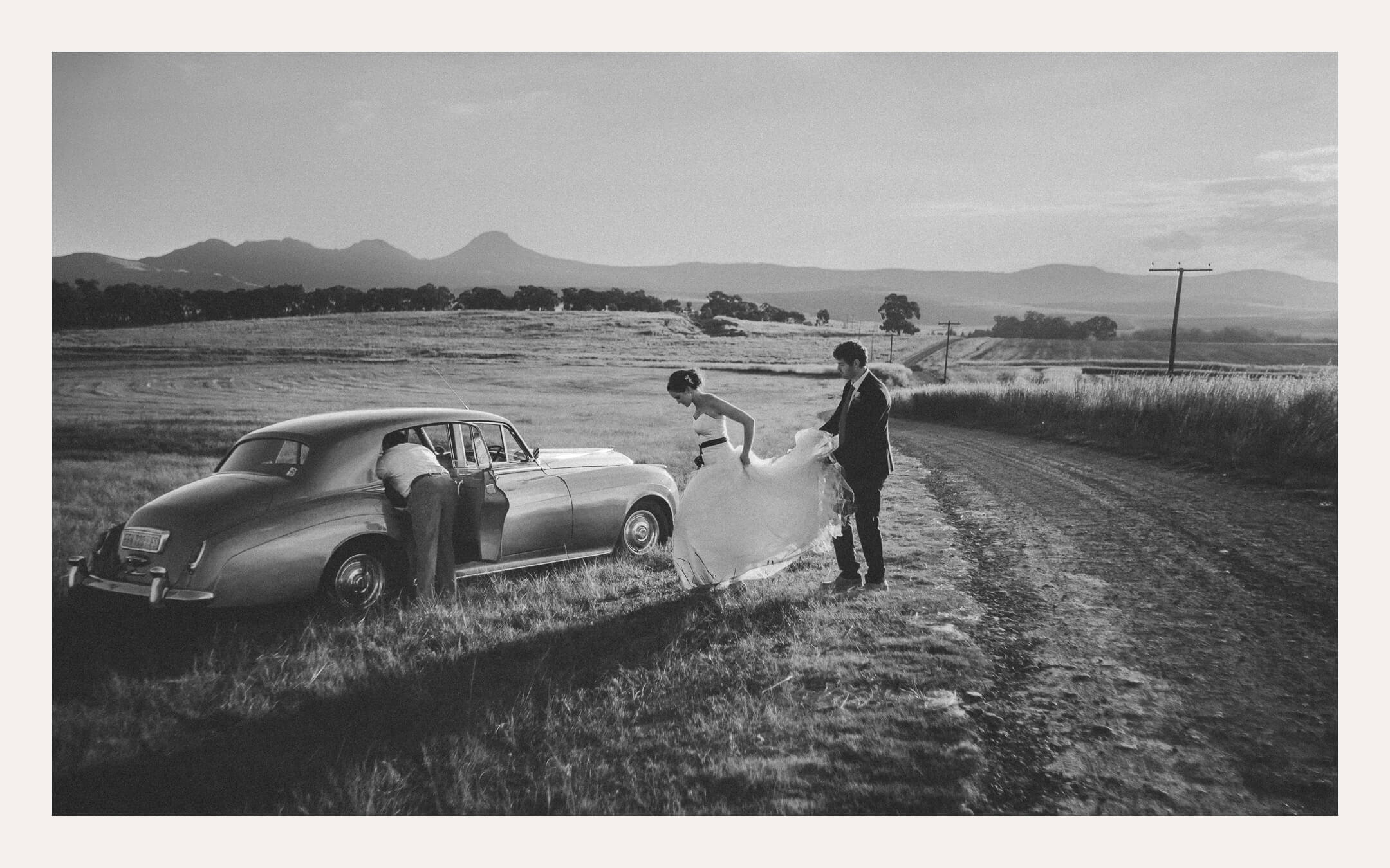 Bride and groom beside a vintage car on a dirt farm road below distant mountains. Ben Viljoen Photography