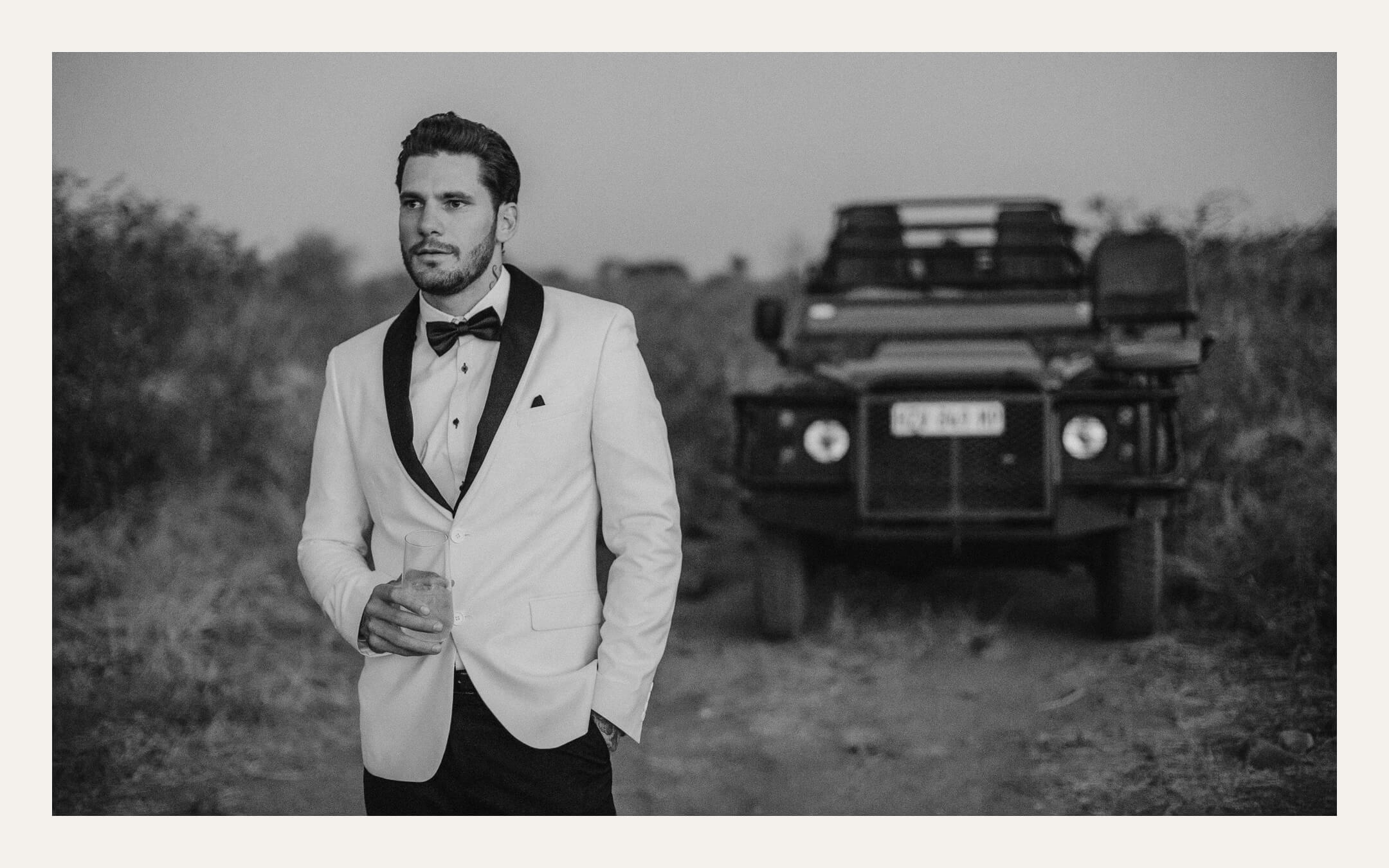 Groom in a white dinner jacket standing in front of a safari vehicle in the bush. Ben Viljoen Photography